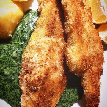 On a white plate are two pieces of pan fried fish on top of creamed nettle sauce. Two boiled potatoes and two lemon wedges next to it.