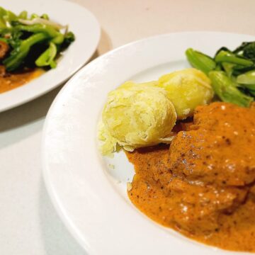 two plates of pork pepper steak in a tomato cream sauce with boiled potatoes and steamed vegetables on a white kitchen bench