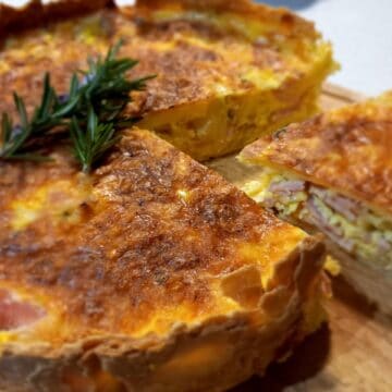 Bacon and egg pie on a wooden chopping board topped with fresh rosemary. A large portion has been cut from the pie showing what it looks like inside.
