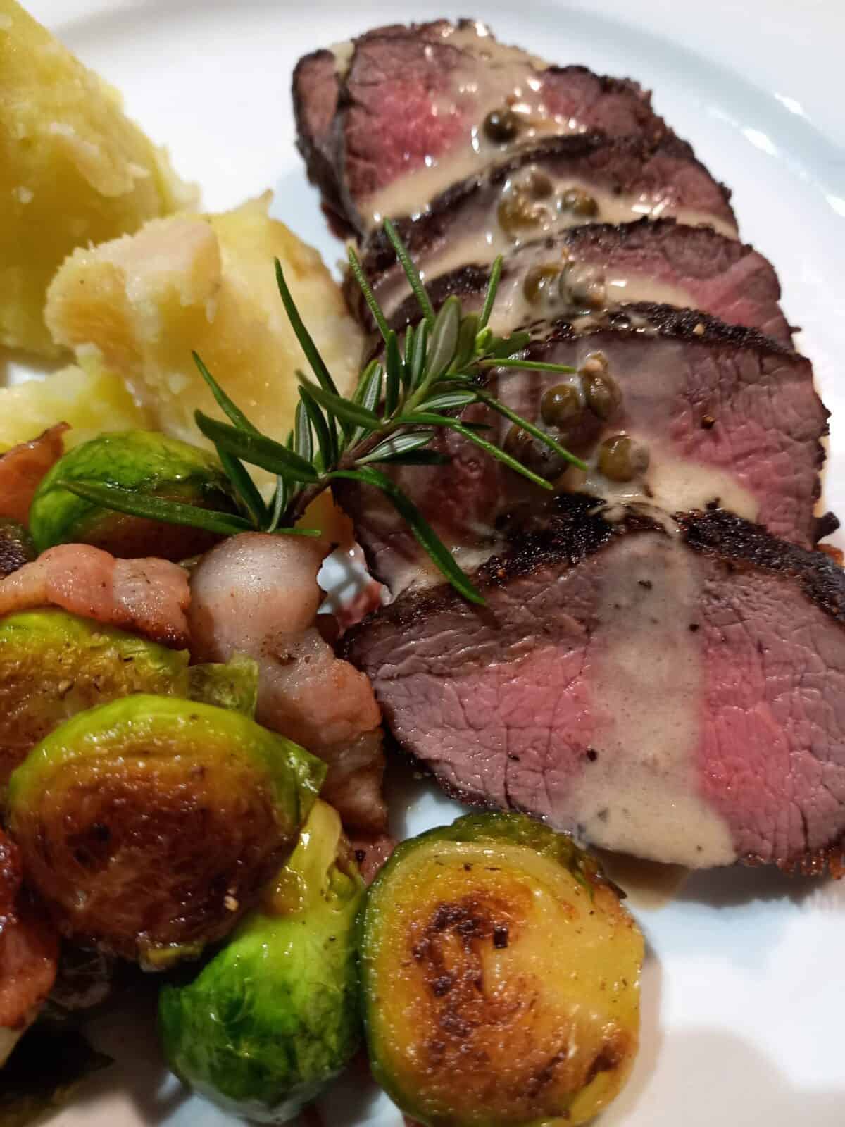 venison meat cooked and sliced with creamy peppercorn sauce. Boiled potatoes and brussel sprouts on a white plate