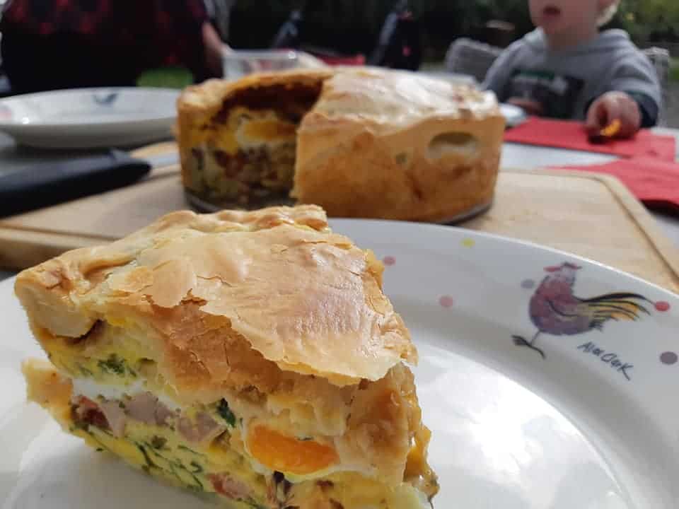 bacon and egg pie slice on a plate. The rest of the pie is on a chopping board in the background