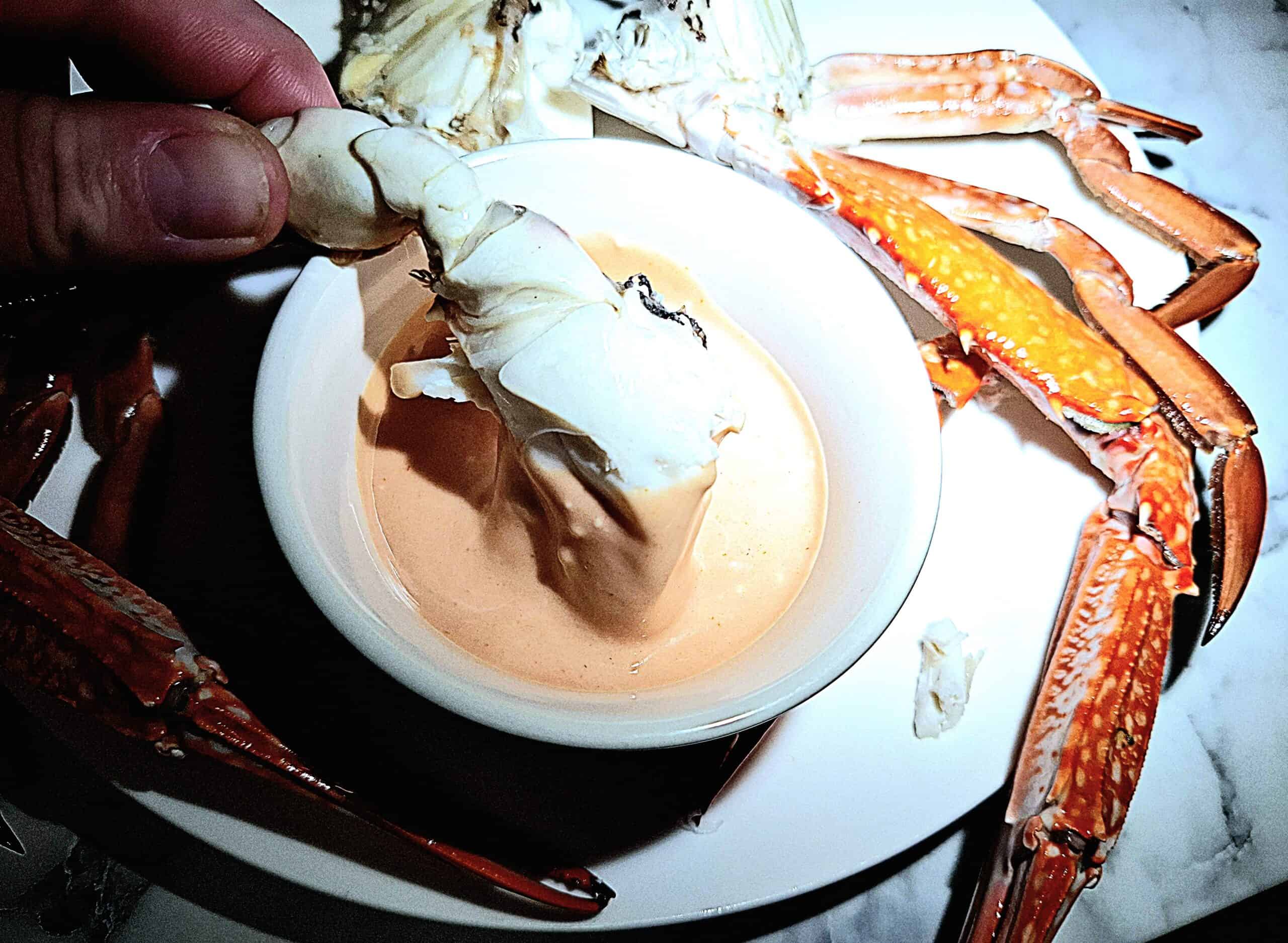 Seafood cocktail sauce in a round ramekin on a white plate with crab pieces on it. A chunk of crab is dipping into the sauce.
