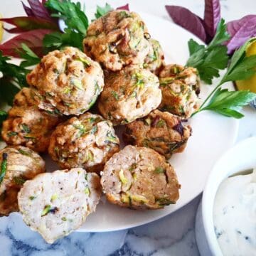 Cooked zucchini balls on a white plate, one cut in half, and next to it a white ramekin with sauce in it. Fresh herbs and lemons next to the plate.