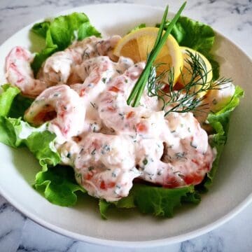 prawn shrimp skagen mix served on top of lettuce leaves with lemon slices, fresh chives and dill on top. Served in a white bowl on a marbled table top