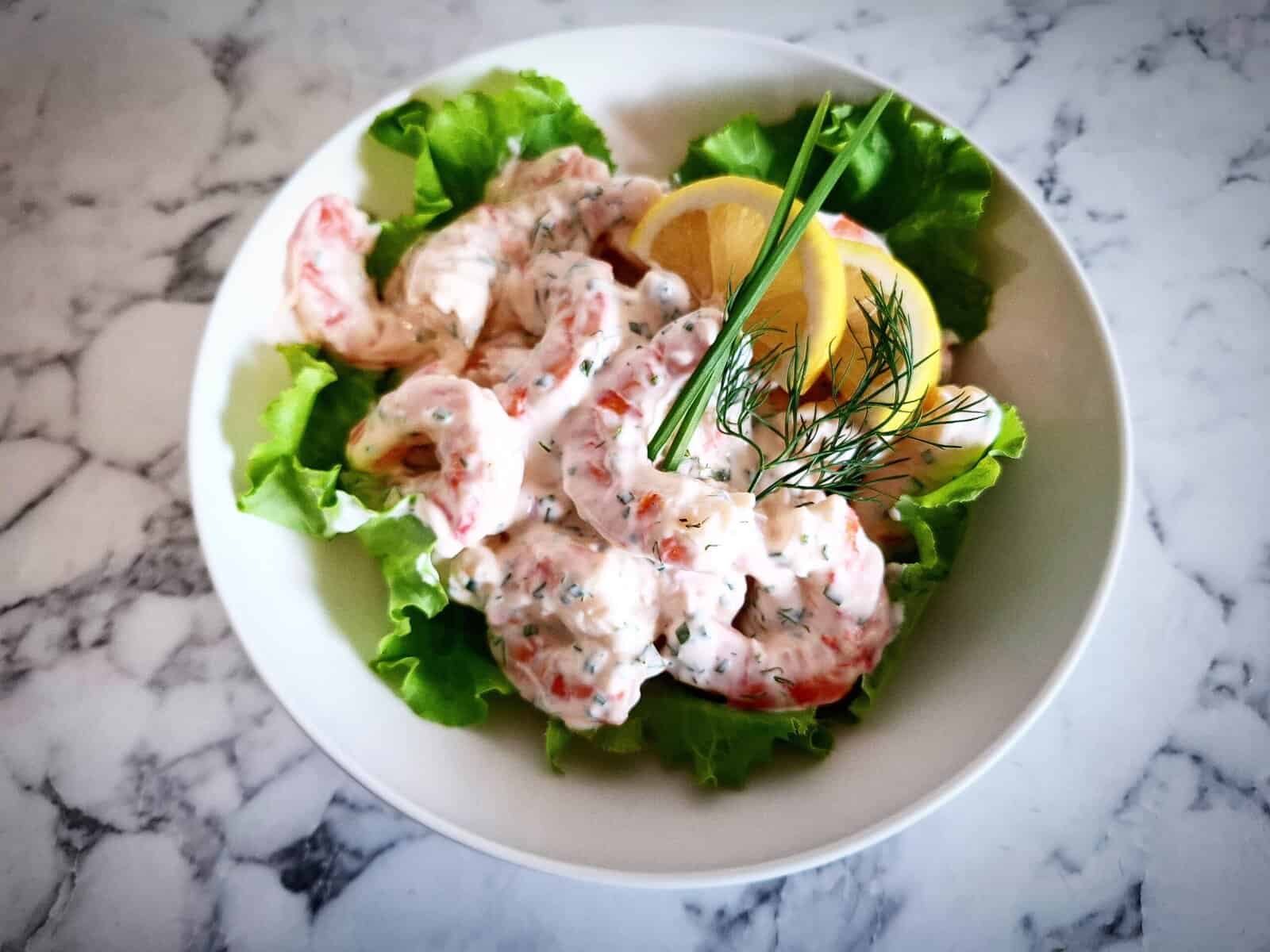 prawn shrimp skagen served on top of lettuce leaves with lemon slices, fresh chives and dill on top. Served in a white bowl on a marbled table top