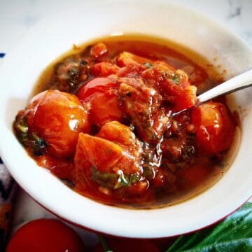 Tomato confit in a ramkein with a spoon in it. Cherry tomatoes and fresh basil leaves below it and a blue, white and yellow tea tale on the left side of it.