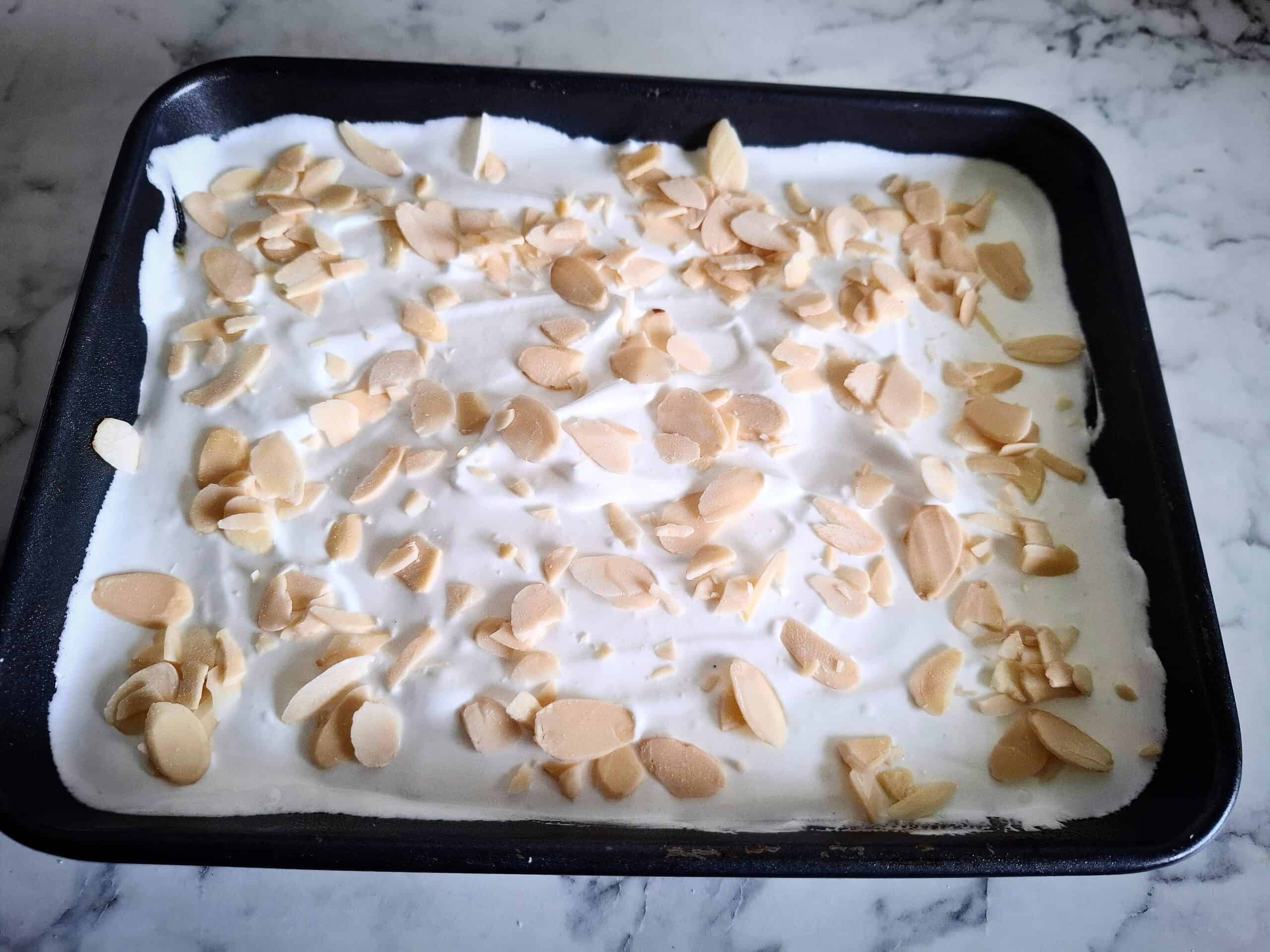 Cake batter has been covered with whipped egg whites and sliced almonds in a black rectangular baking tin on a marbled bench top.