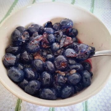 Macerated blueberries in a white, round bowl with a spoon in it on a white and green striped tea towel.