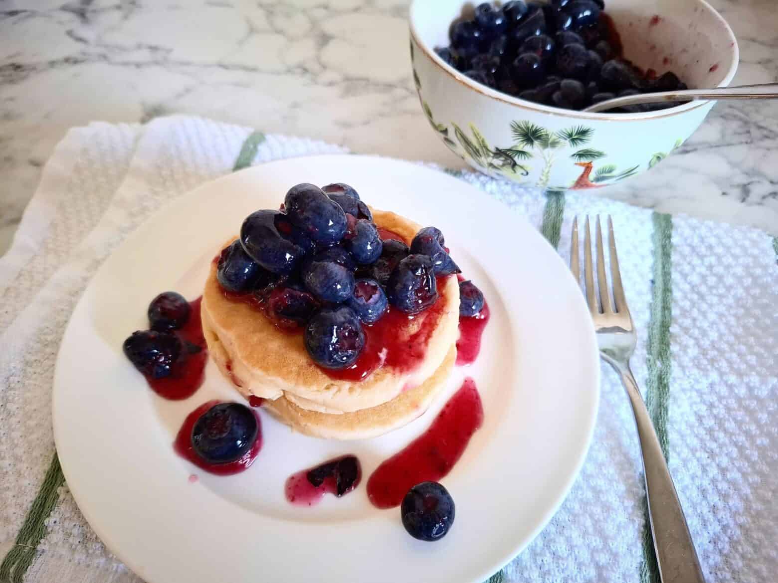 Three stacked pikelets with macerated blueberries on top on a white round plate. A fork next to it on a white and green striped tea towel. A safari printed serving bowl half filled with macerated blueberries and a spoon behind the pikelet plate.
