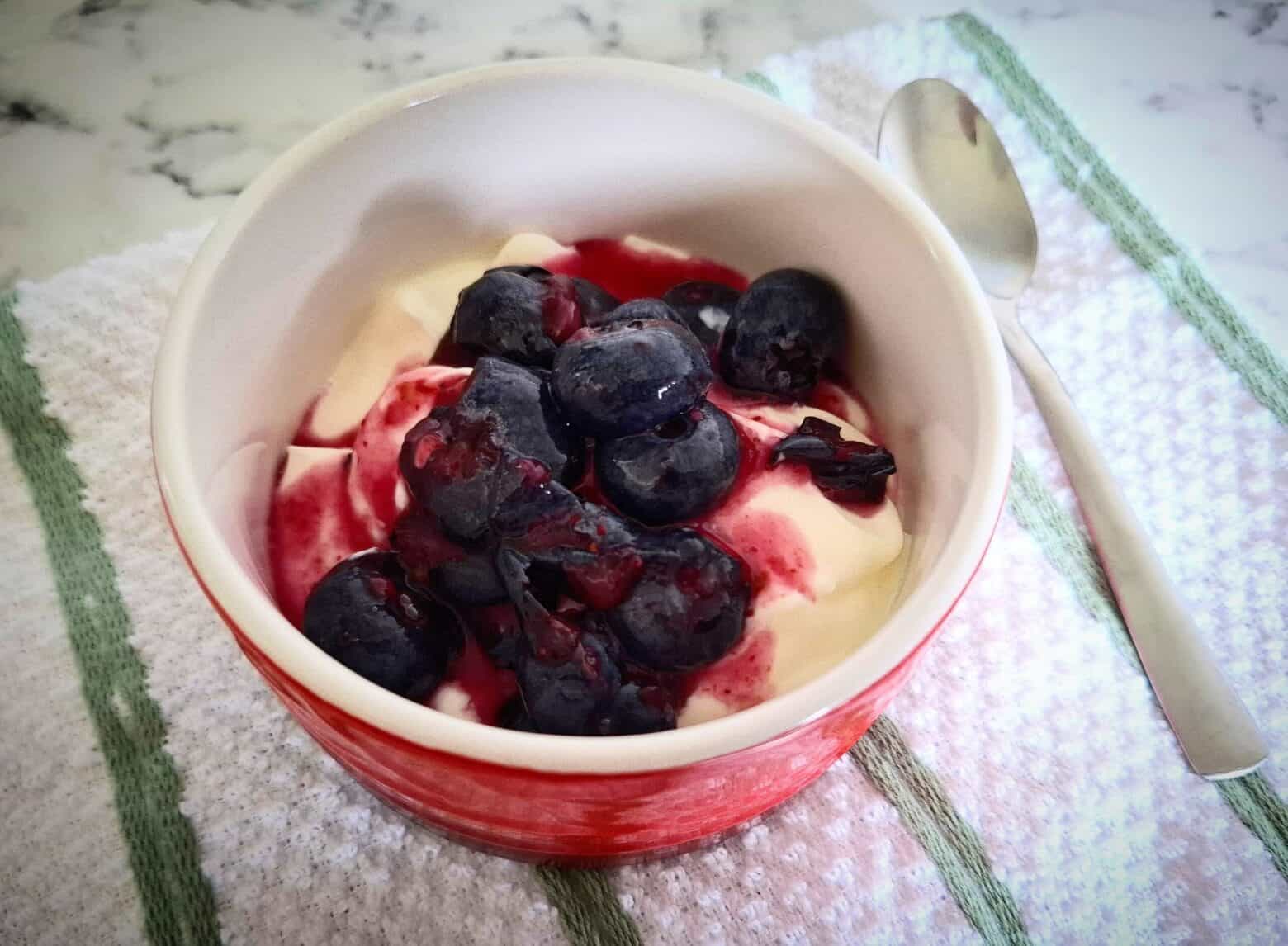Custard with macerated blueberries on top in a red and white ramekin with a spoon next to it on a white and green striped tea towel.