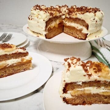 A banoffee cake with two portions cut out of it and served on two dessert plates in front of it. Two plain dessert plates and two forks behind the cake portions. A green and white striped tea towel with cake serving cutlery on top on the right hand side next to the cake.