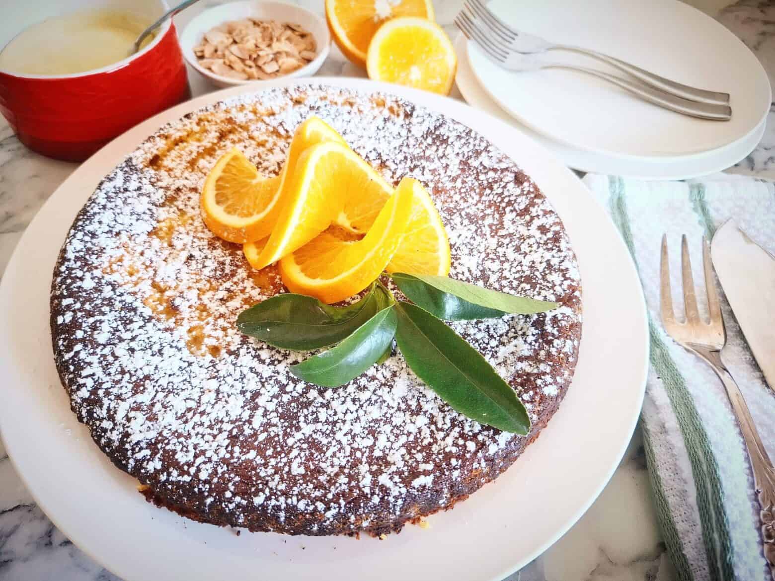 An orange and almond cake dusted with icing sugar and garnished with three swirly orange slices and a small bunch of orange leaves in the center of the cake. Behind the cake is a red ramekin of icing, a white ramekin of roasted almond slices, two orange halves and two white dessert plates with two forks on top. On the right hand side of the cake is a green and white striped tea towel with cake serving cutlery on top.
