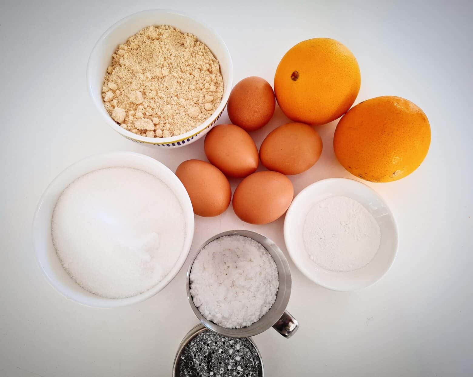 Ingredients for orange and almond cake in ramekins on a white table top.
