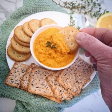 Carrot hummus served in a white ramekin topped with two thyme leaves on a white plate. A hand is picking up carrot hummus with a round cracker. On the plate are seven round crackers, five wholegrain, rectangular crackers and a spoon. There is a green tea towel underneath the plate, half a lemon and fresh thyme leaves in the background.
