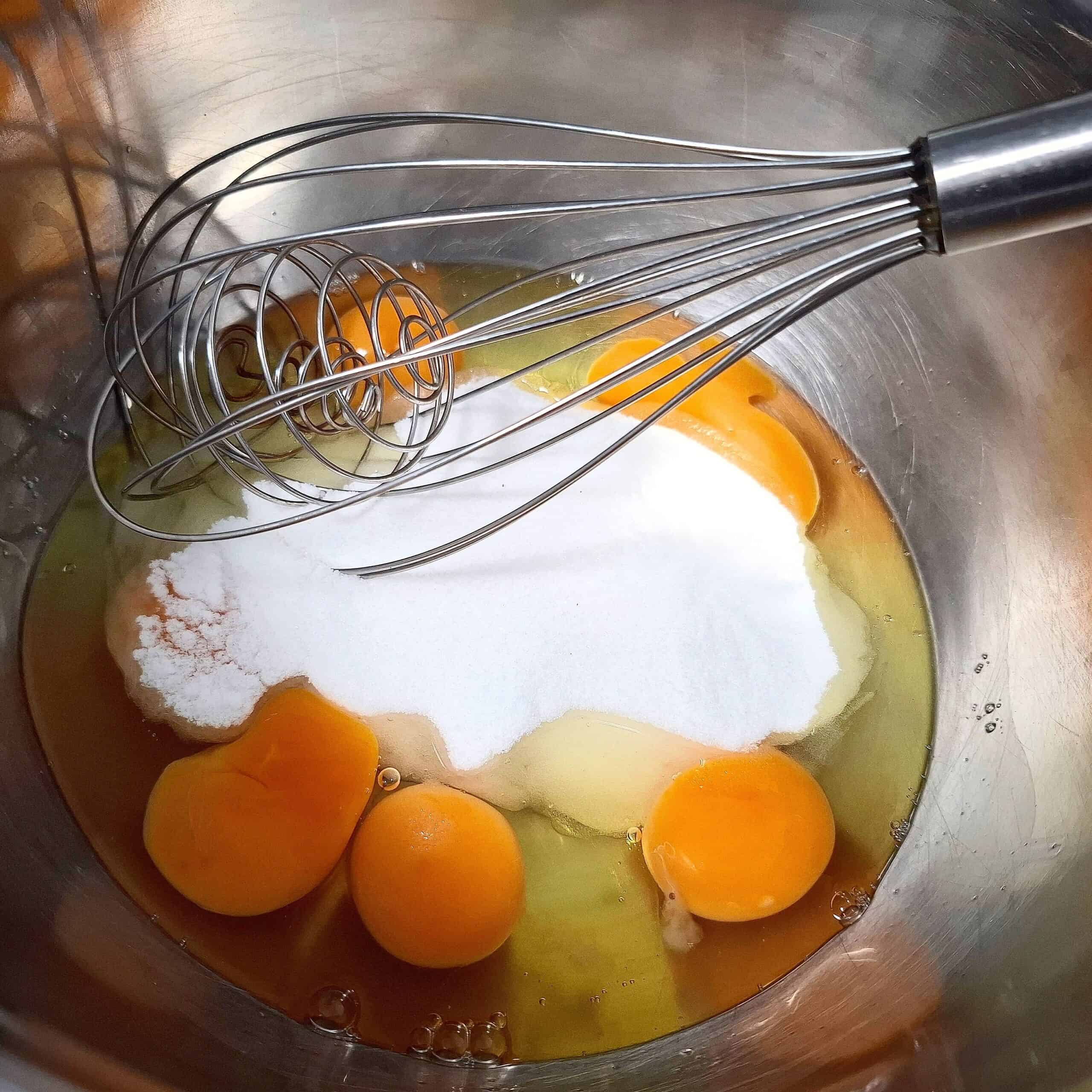 Eggs and sugar in a stainless steel bowl with a balloon whisk resting in it.