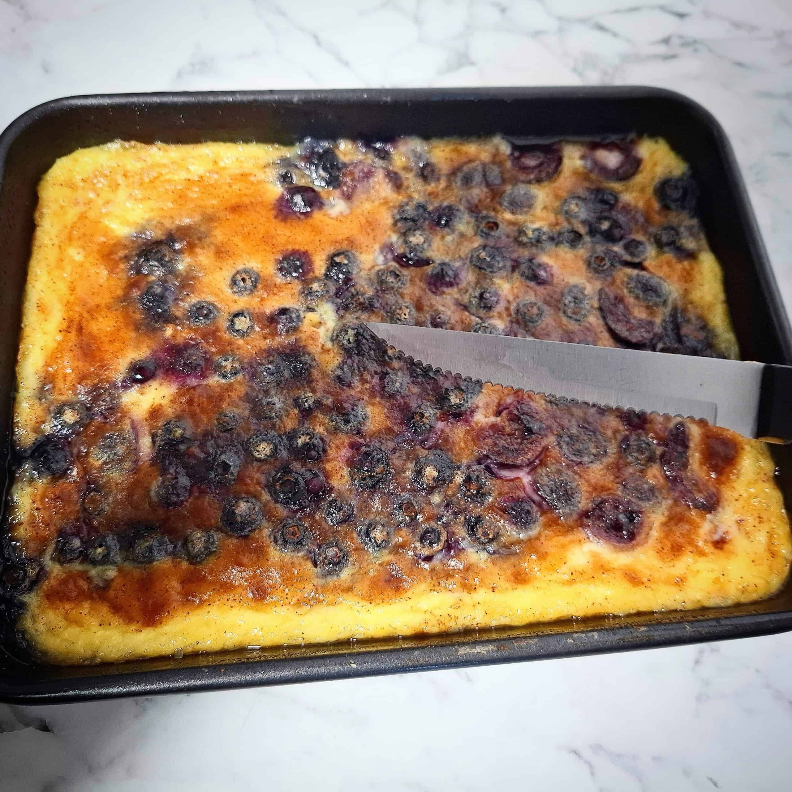 Blueberry baked custard in a black rectangular baking tray on a marbled bench top. A clean knife bade above it.