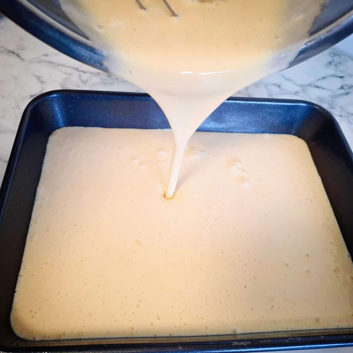 Pouring raw custard mixture into a rectangular black baking tray from a stainless steel bowl.
