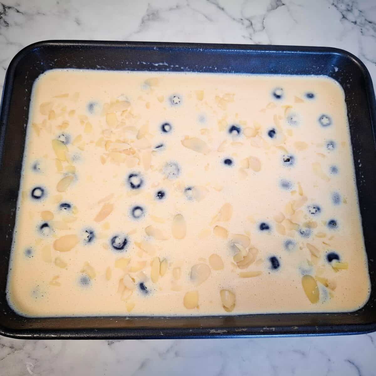 Raw blueberry custard mixture in a rectangular baking tray with sliced almonds on top.
