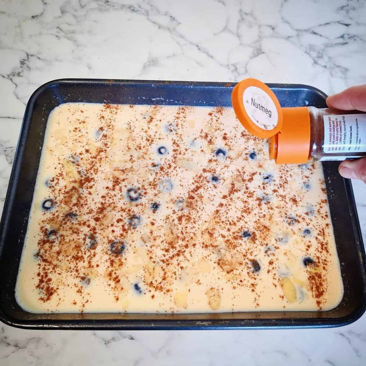 Dusting nutmeg on top of the raw blueberry custard mixture from a shaker with an orange lid