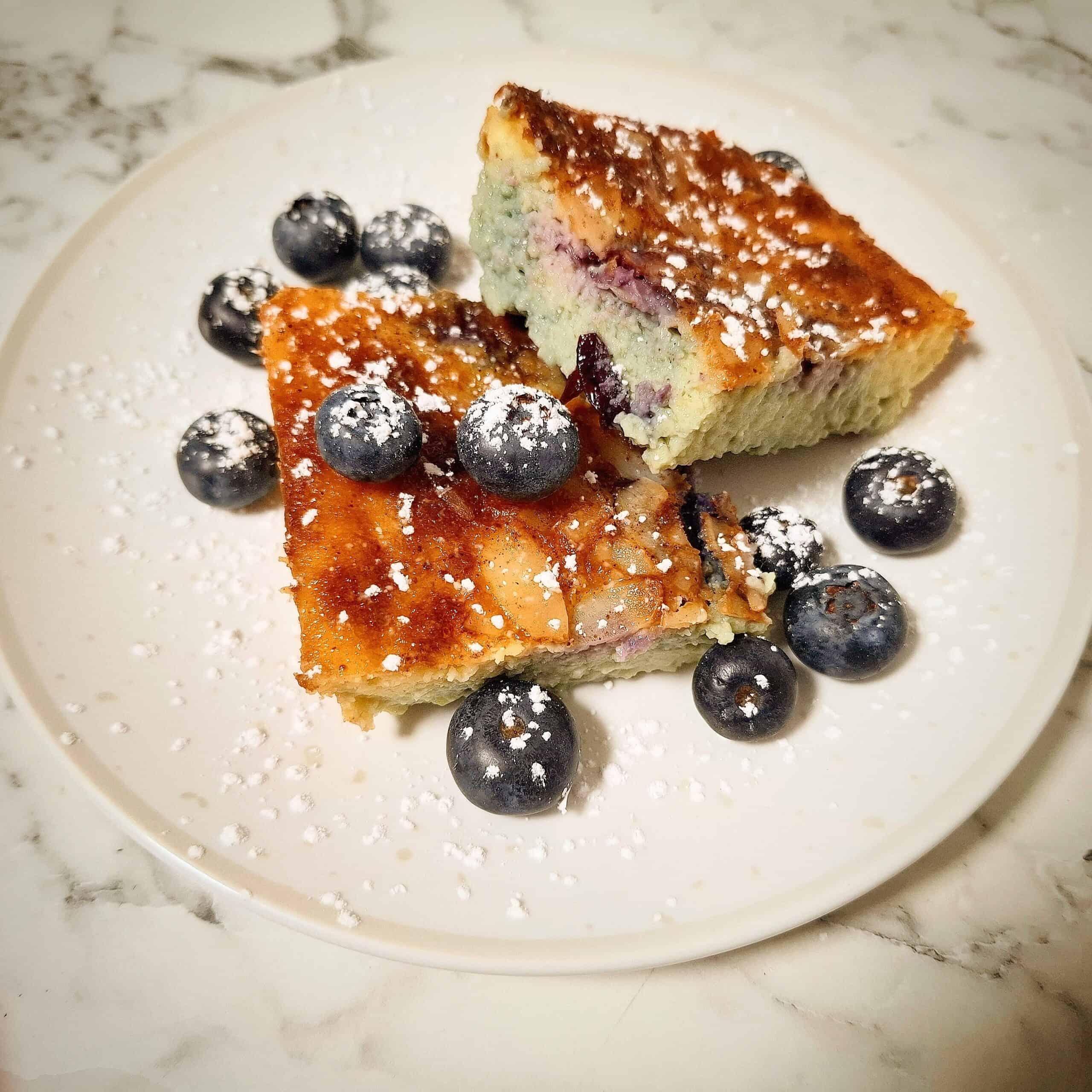 Two pieces of Blueberry baked custard and fresh blueberries dusted with icing sugar on a white dessert plate.