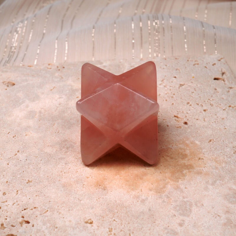 Natural Rose Quartz Merkaba ornament displayed on a light stone surface, with soft pink tones and a semi-translucent finish.