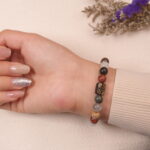 A hand wearing the multicolor crystal bracelet with a black and gold floral charm, showcasing the colorful beads including Tiger Eye and Agate. The bracelet enhances the natural tones of the skin and adds a touch of sophistication.