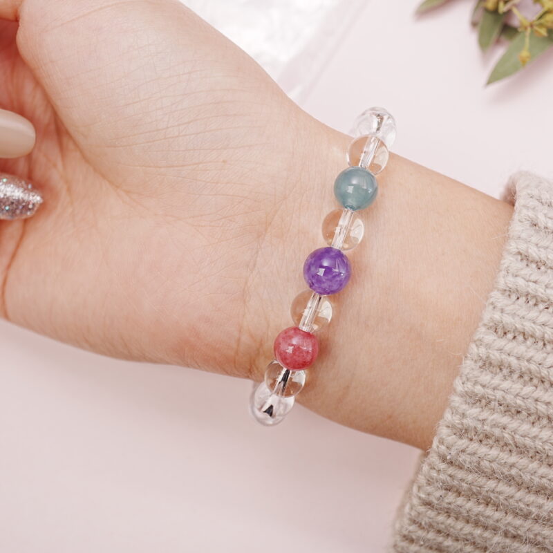 Model wearing a clear quartz bracelet featuring red, purple, and teal gemstone beads