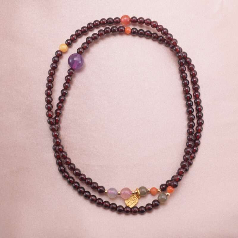 A beaded necklace made of deep red garnet with amethyst, yellow crystal, and red agate accent beads displayed on a soft background.