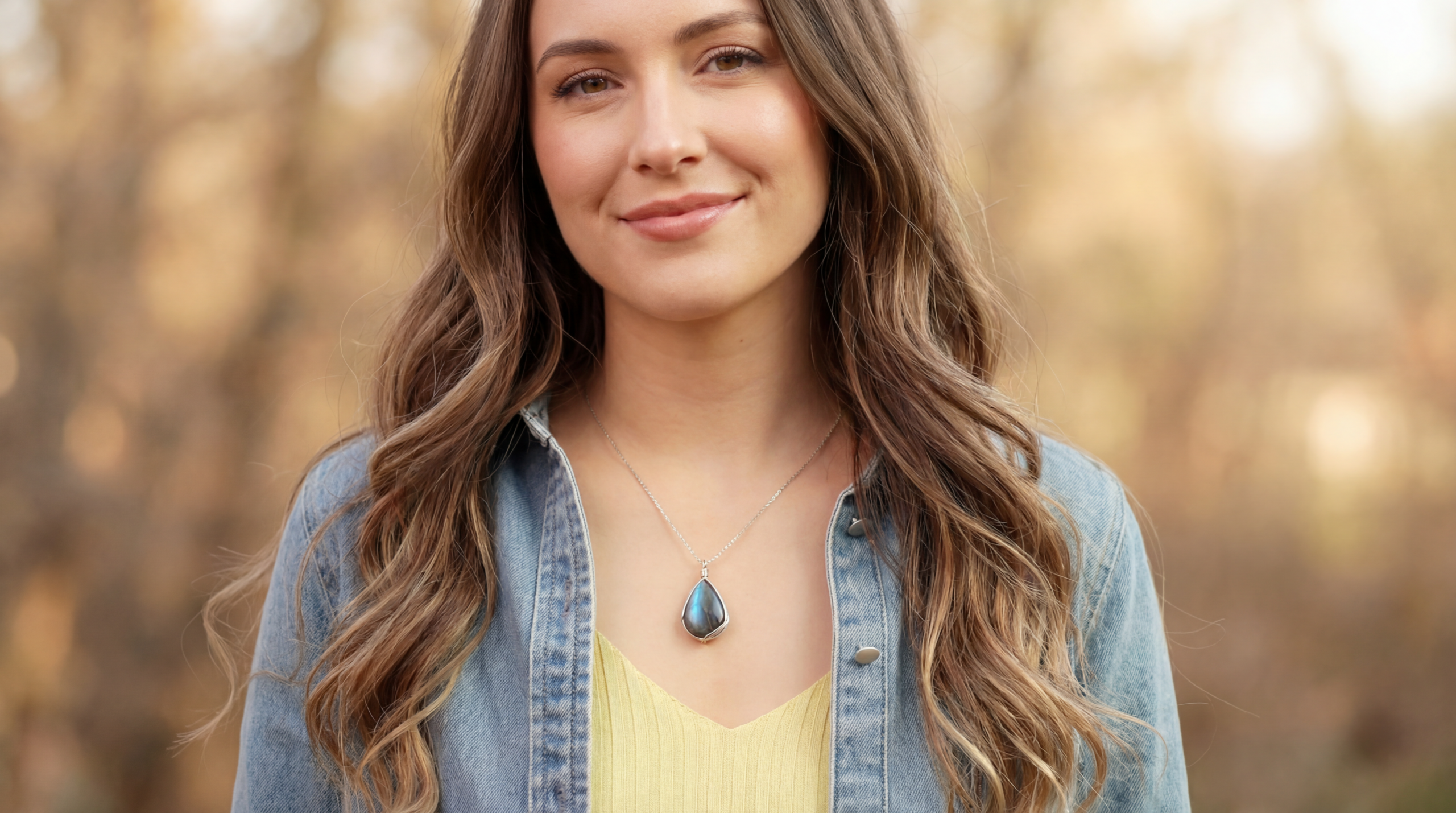 A woman wearing a blue gemstone pendant necklace outdoors in warm natural light, showcasing elegant everyday jewelry styling with a soft blurred background.