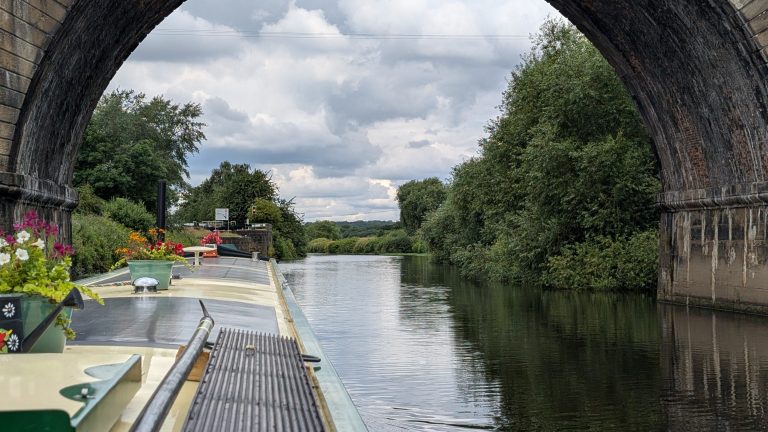 Narrowboat cruising through a scenic canal under a stone bridge in the UK.