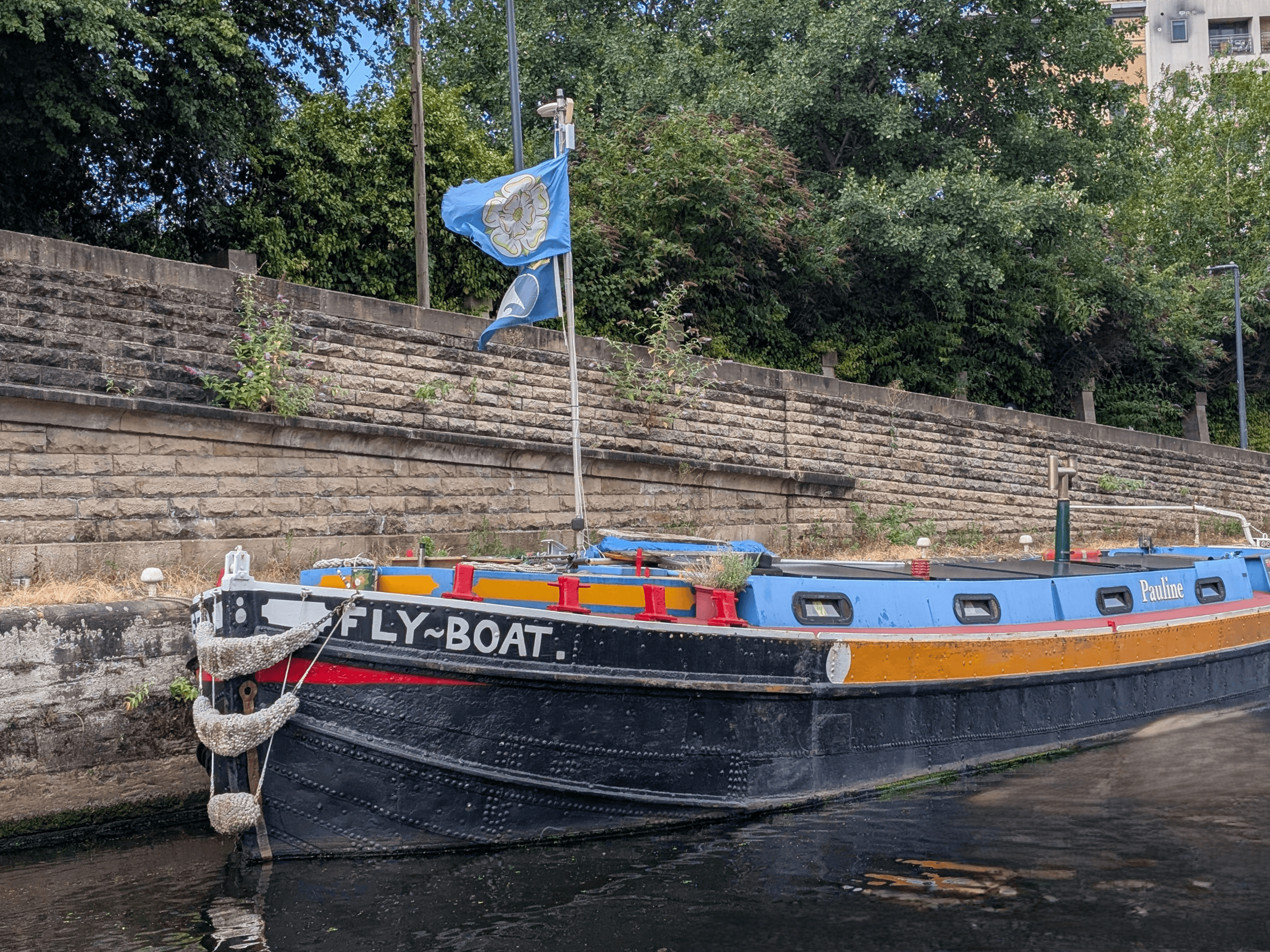 Traditional narrowboat moored along a canal with a blue flag and lush greenery in the background.