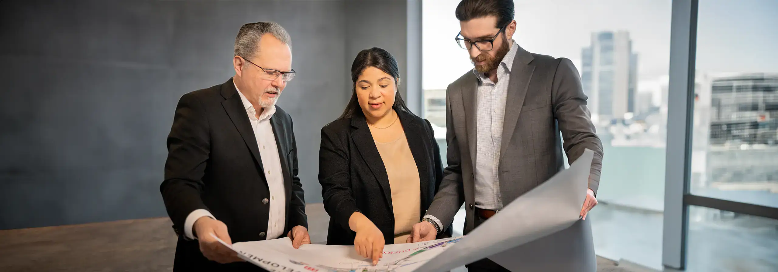 Three professionals reviewing blueprints in a modern office with city view.