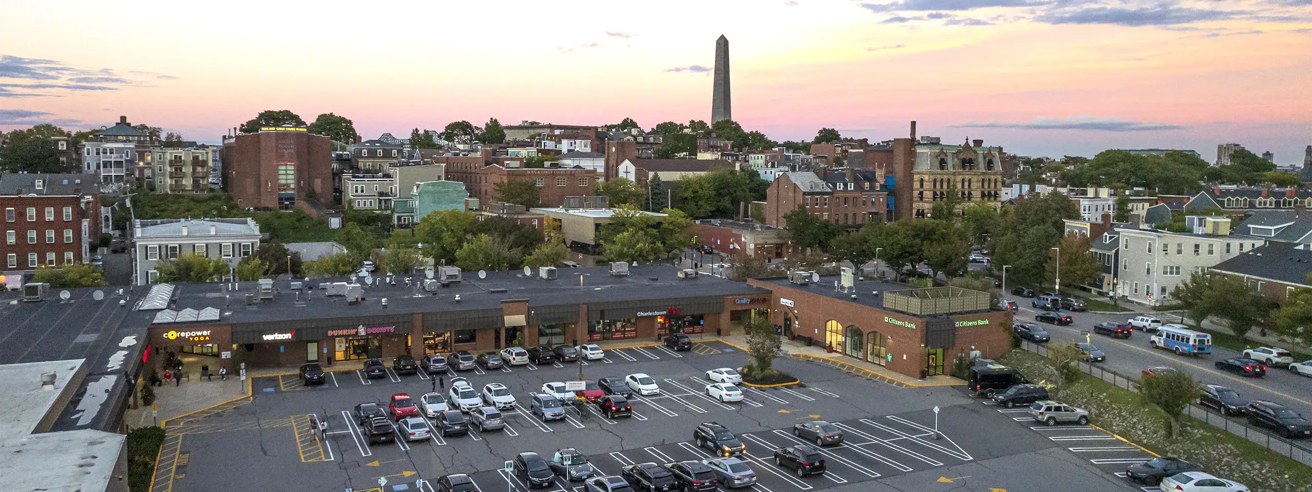 Aerial view of a shopping center parking lot with city skyline in background at sunset.