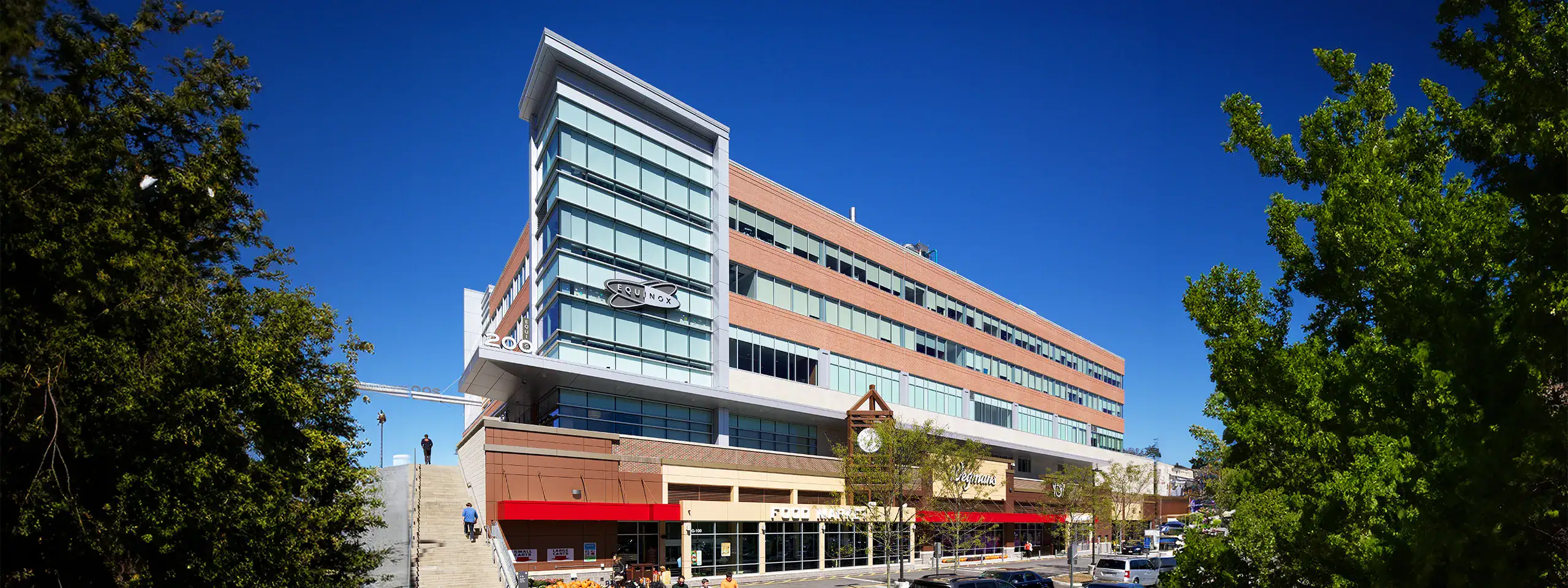 Modern multi-story building with glass facade and red awnings, surrounded by trees.