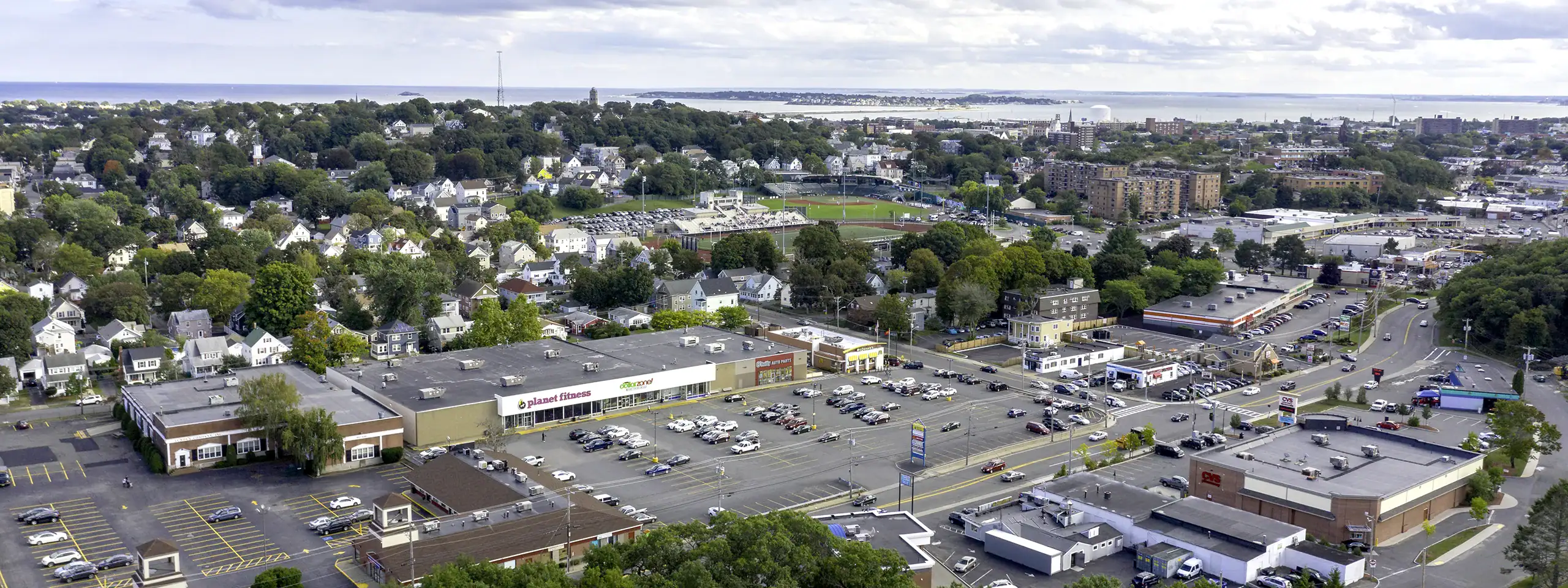 Aerial view of a shopping center with parking lot and surrounding buildings in New England.
