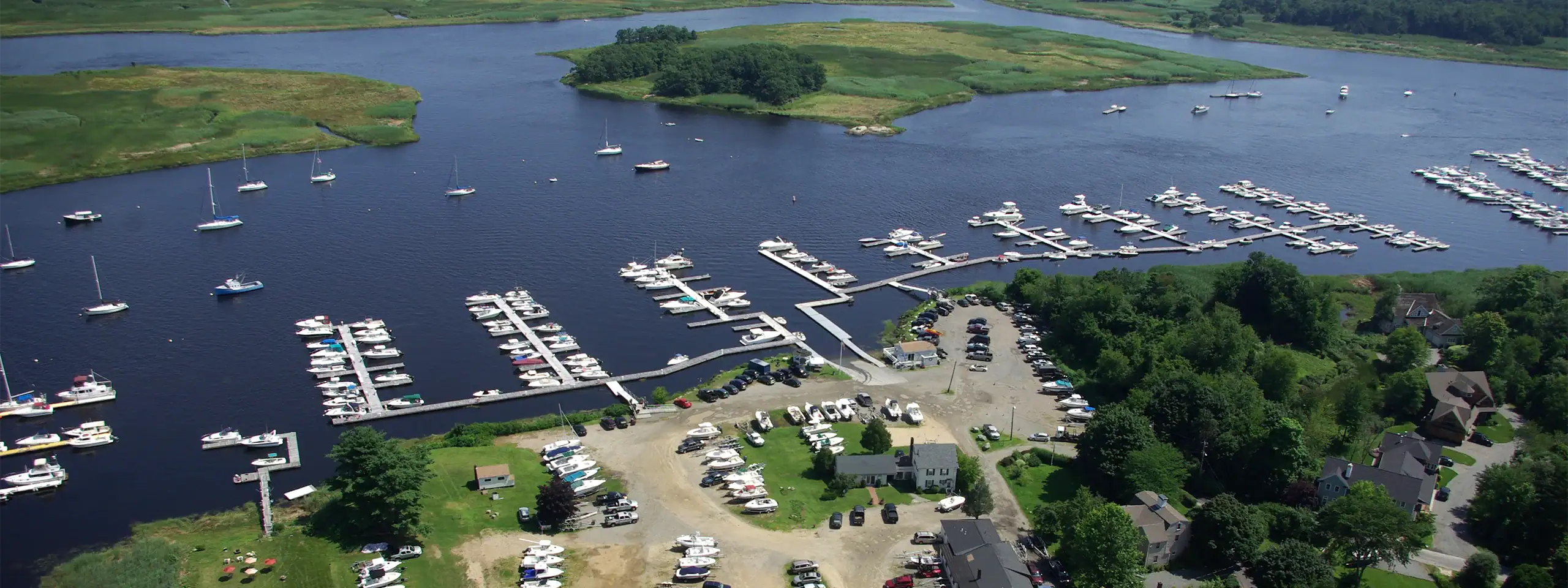 Aerial view of a marina with boats docked on a river surrounded by green trees and land.