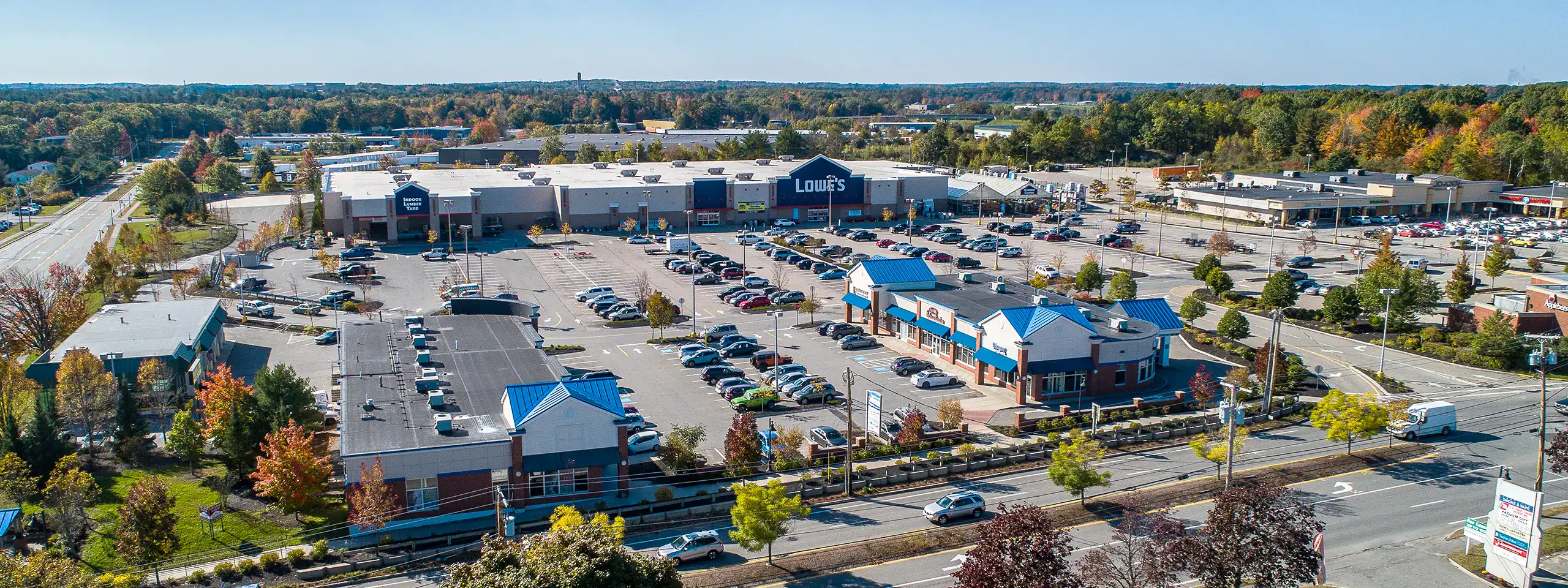 Aerial view of a shopping center parking lot with stores and trees in fall colors.