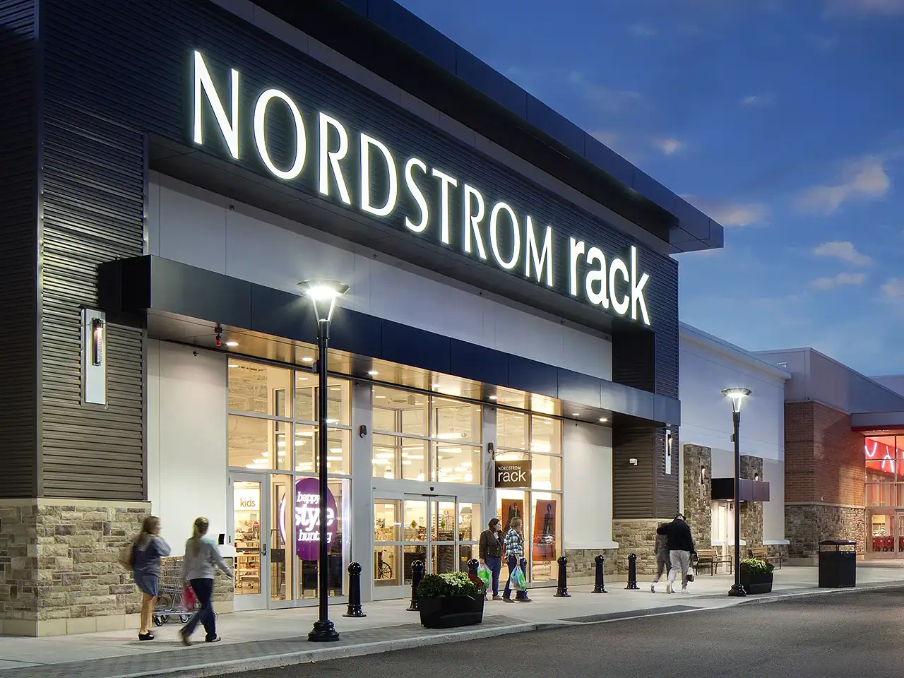 Exterior view of Nordstrom Rack store at dusk with shoppers entering.
