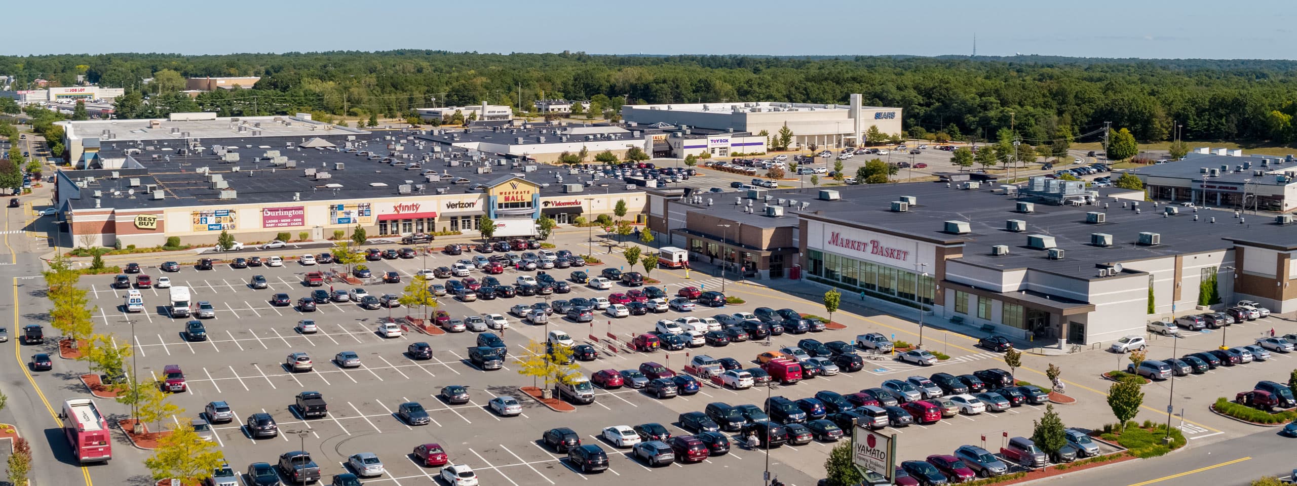 Large shopping center parking lot with numerous cars and retail stores in the background.