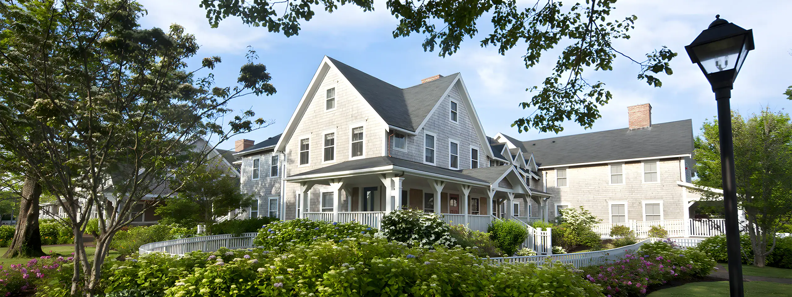 Historic gray building with white trim and a porch, surrounded by trees and flowers.