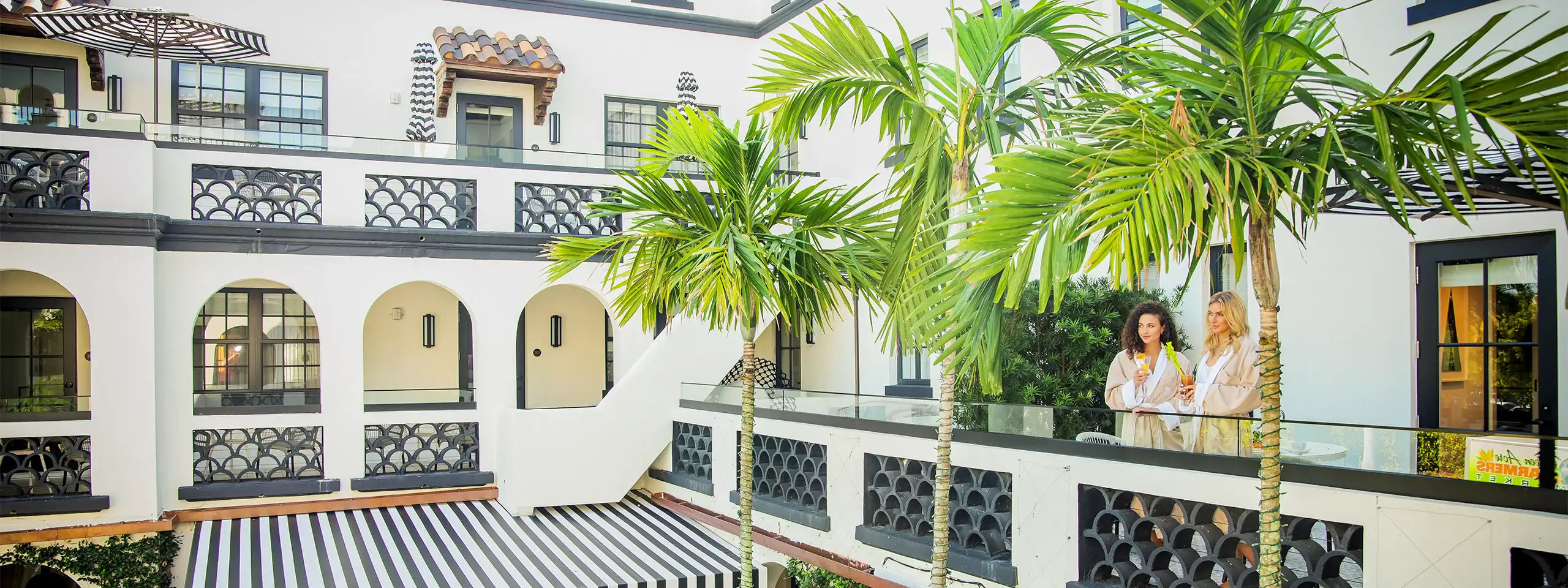Two women talking on a balcony with palm trees and white building in the background.