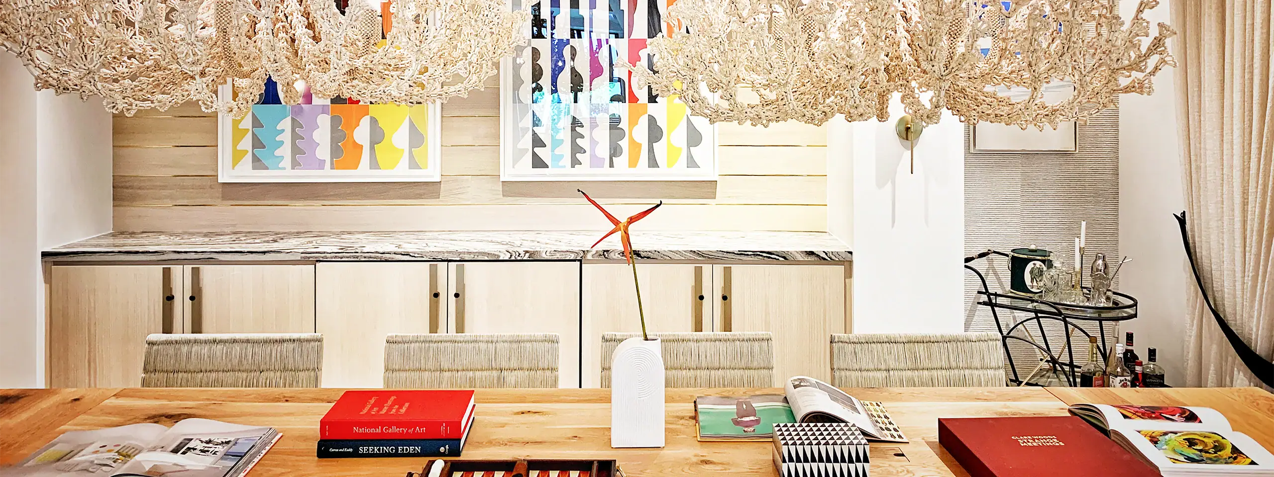 Modern dining room with wooden table, colorful artwork, and elegant chandeliers.