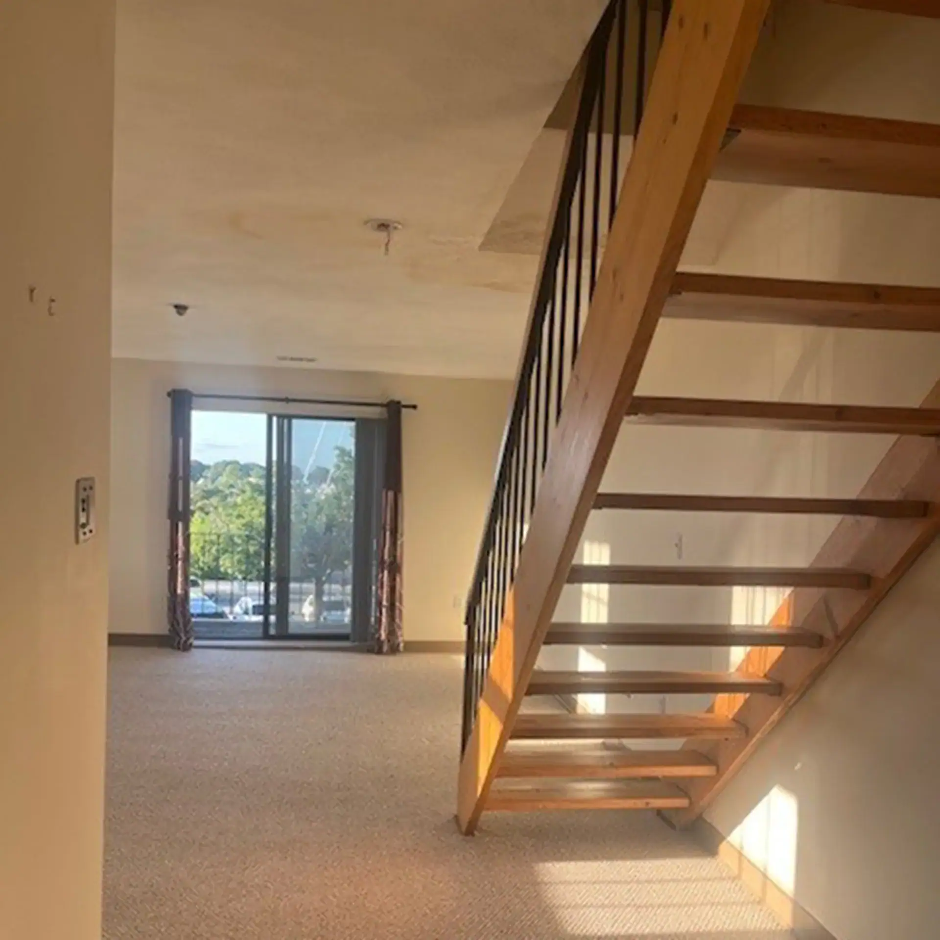 Empty living room with staircase and sliding glass door leading outside.