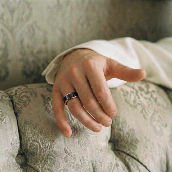 Relaxing on a sofa, you can just see a mans arm and hand. Wearing a white shirt, he is wearing a tungsten ring on this wedding finger. It is a black and rose gold ring.