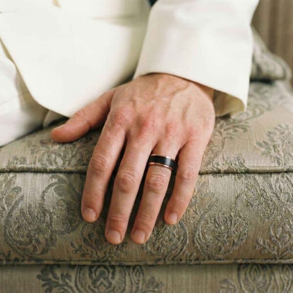 Sitting on a sofa, this is a close up image of a mans hand. You can see that the man is wearing a white suit. The ring is a black and rose gold band made with tungsten.