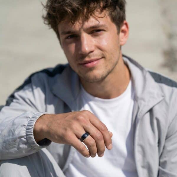 Sitting in a relaxed pose and wearing a hoodie, a man smiles into the camera. He rests his hand on his knee so that there is a prominent view of his hand. He is wearing a black and silver domed ring made with tungsten.