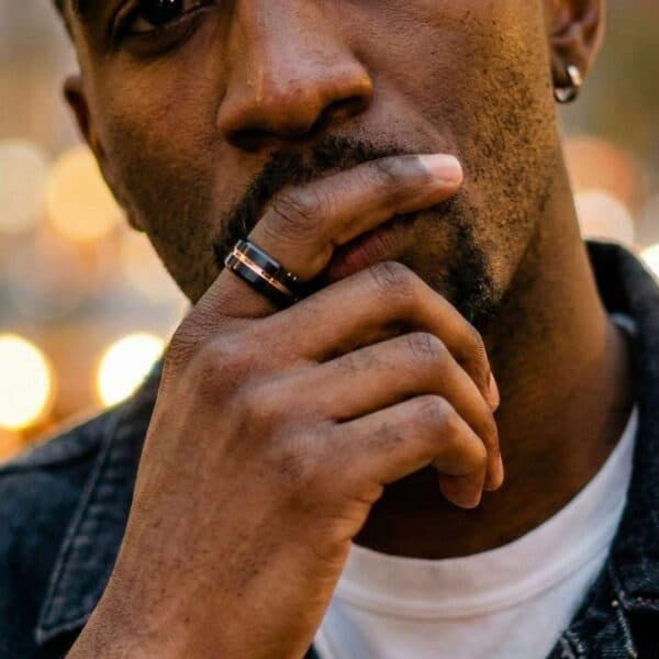 A close up of a mans face. Holding his hand in a thoughtful pose across his mouth, he is wearing a black tungsten ring. It features a groove of rose gold.