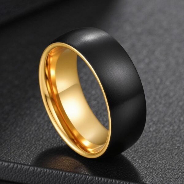 On a smooth black leather surface, a tungsten ring stands balanced in an upright position. It is a black ring with a brushed finish, and a gold interior.