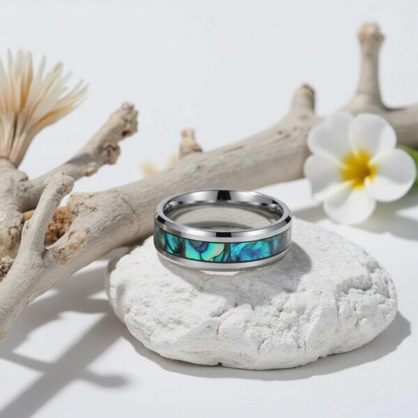 A professional product photo shoot of a mens ring. The ring is set on a piece of white chalk. There is a white flower and a white piece of wood. The ring is a silver tungsten ring with an inlay of abalone shell.