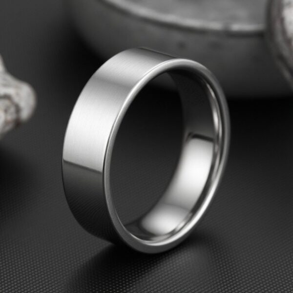 With a grey minimalist background, a ring stands upright. It is a silver tungsten ring that is flat.